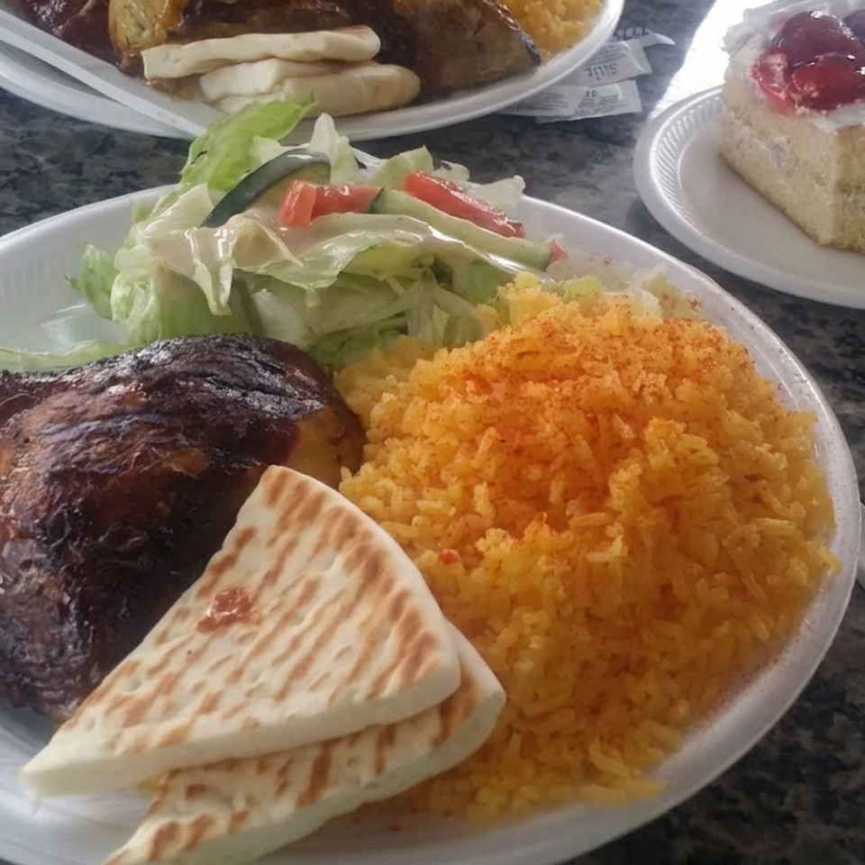 Plate of grilled chicken, yellow rice, salad, and flatbread, with cake and another meal on a granite table.